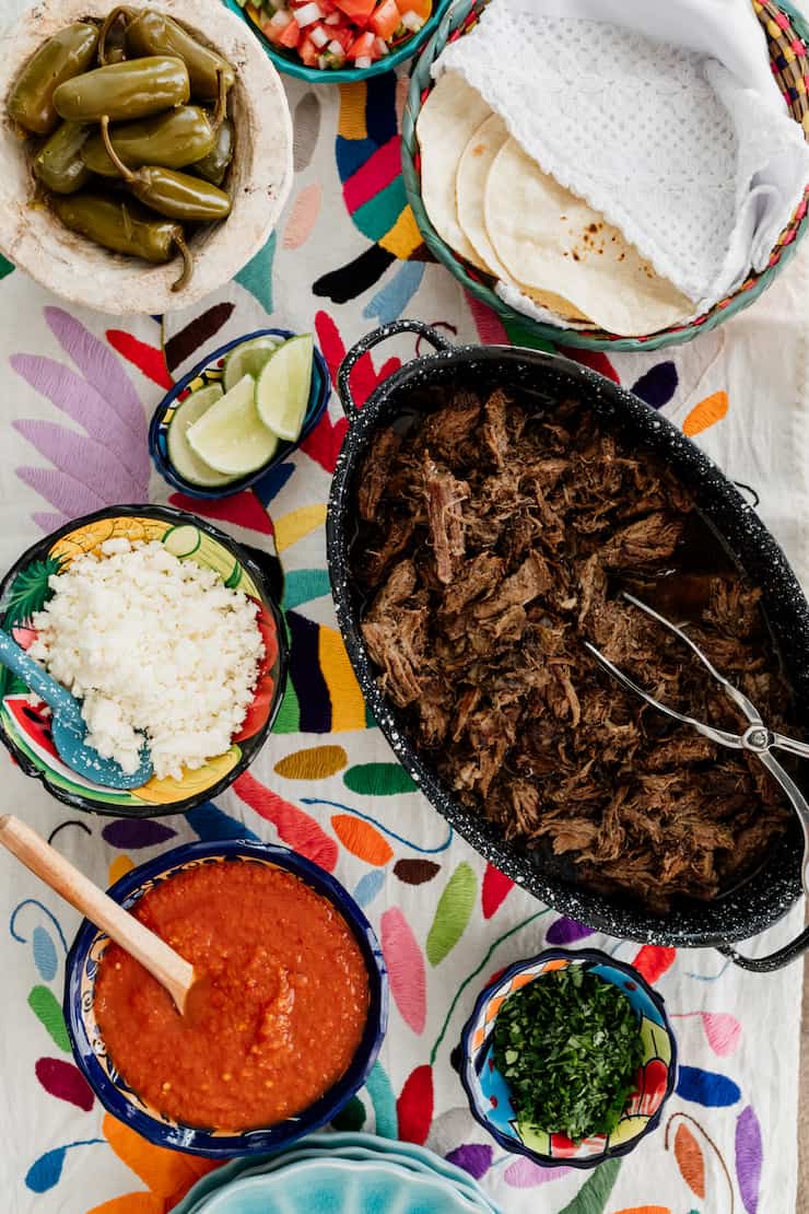 how to set up a serve yourself taco bar with tortillas, suadero style meat, onions, salsa, cilantro and lime wedges on a colorful runner 