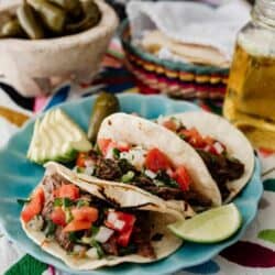 three slow cooker brisket tacos on a turquoise plate topped with pico de gallo and carne deshebrada