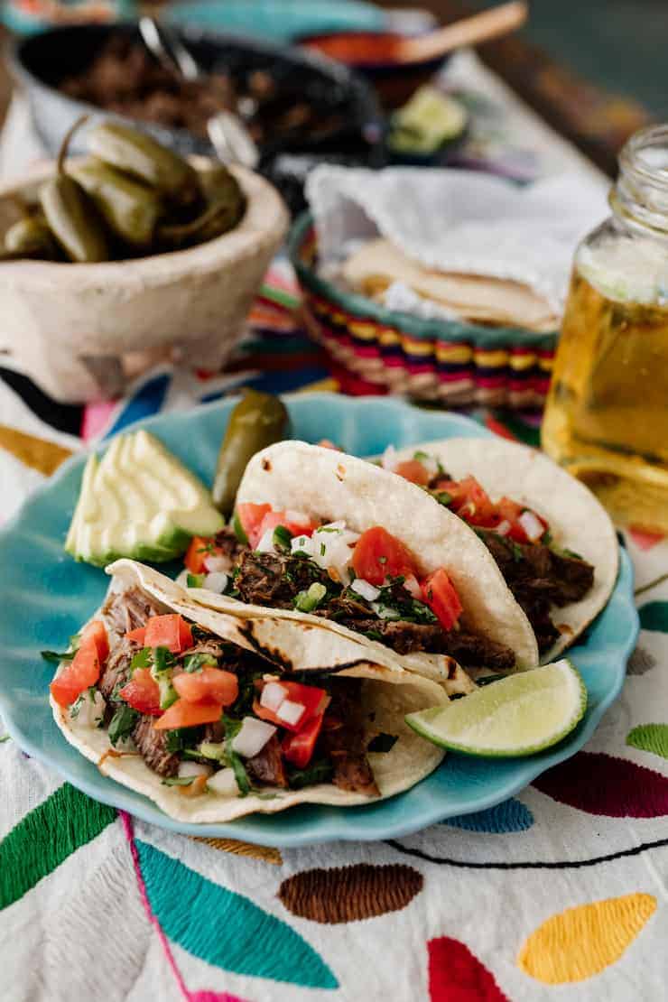 three slow cooker brisket tacos Suadero on a turquoise plate topped with pico de gallo and with a wedge of lime, sliced avocado and escabeche on the side