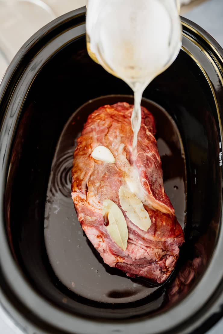 beer being poured into the bowl of a crockpot with brisket, bay leaves and garlic