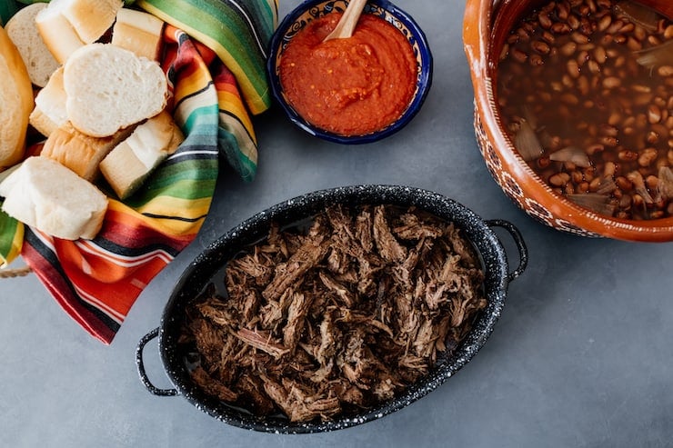 shredded crockpot brisket in a handled black serving bowl with a blue bowl of red salsa, a brown bowl of frijoles de la olla and a striped tea towel lined bread basket with bolillos
