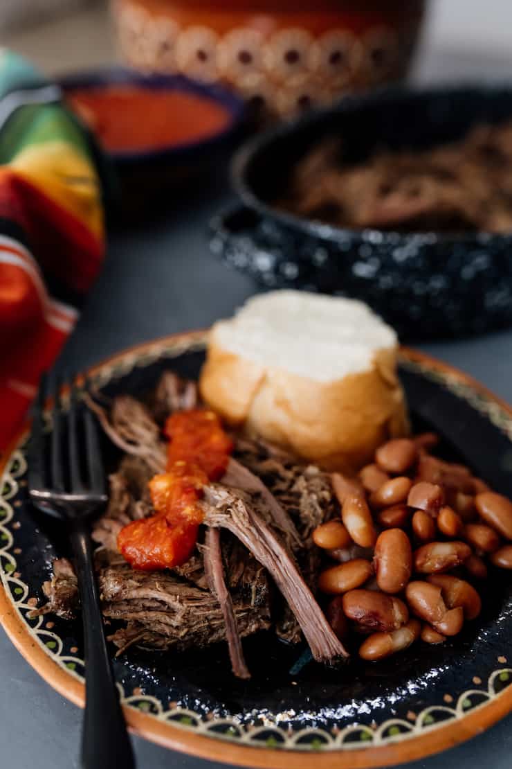 shredded slow cooker beef brisket on a black plate with salsa, beans and a bolillo roll