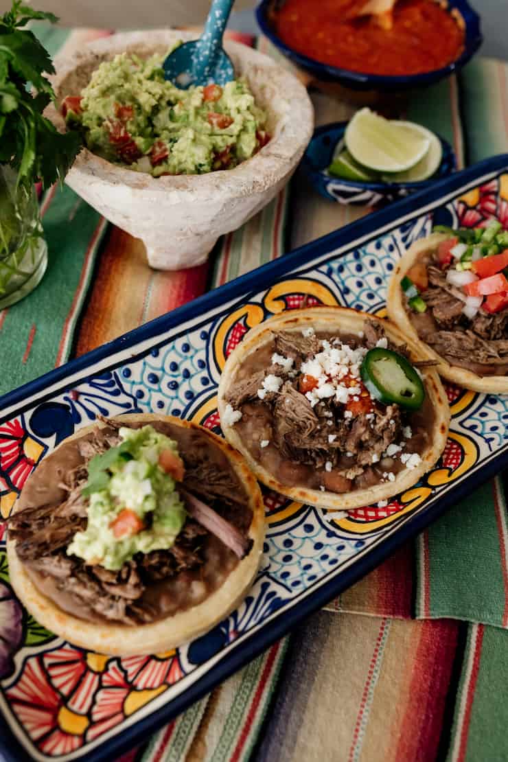 three beef brisket topped mexican sopes on a colorful rectangular serving platter with a bowl of guacamole and salsa in the background 
