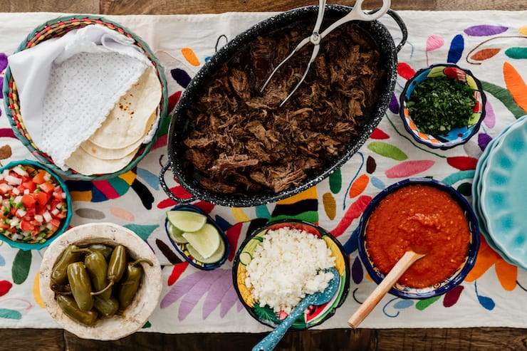 colorful tapestry under a party style taco bar with tortillas, carne deshebrada (suadero), escabeche, salsa, onions and lime wedges