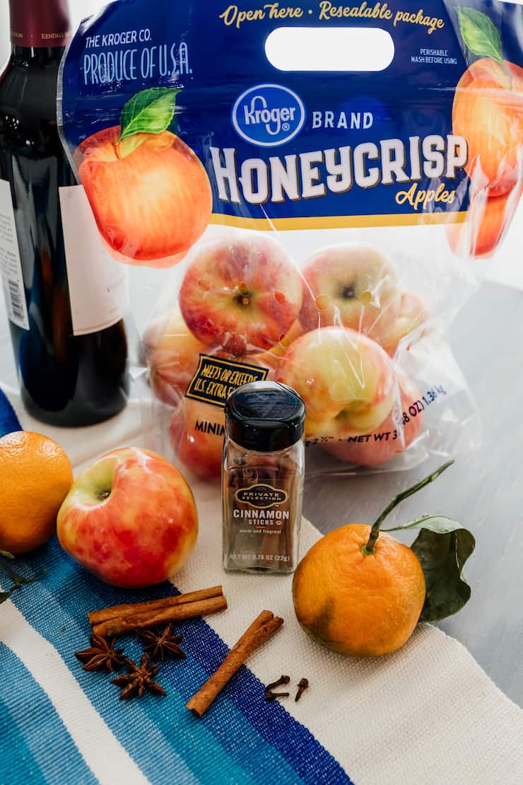 mise en place for mulled wine recipe - bottle of red wine, honeycrisp apples, cinnamon sticks, whole star anise and oranges on the vine