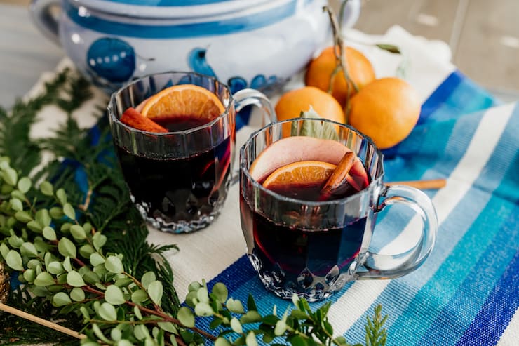 two mugs of spiced mulled wine with cinnamon stick and fruit slice garnishes on a blue and white tablescape with greenery