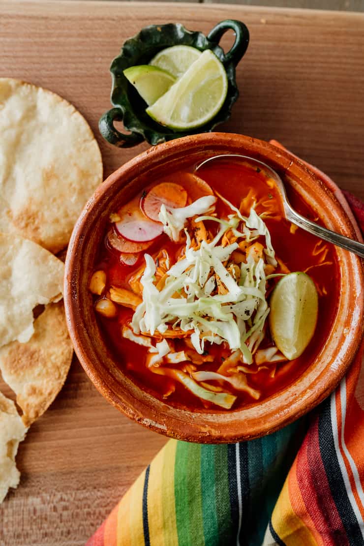 red chicken pozole in a terra cotta bowl with a silver spoon and tortillas to the side