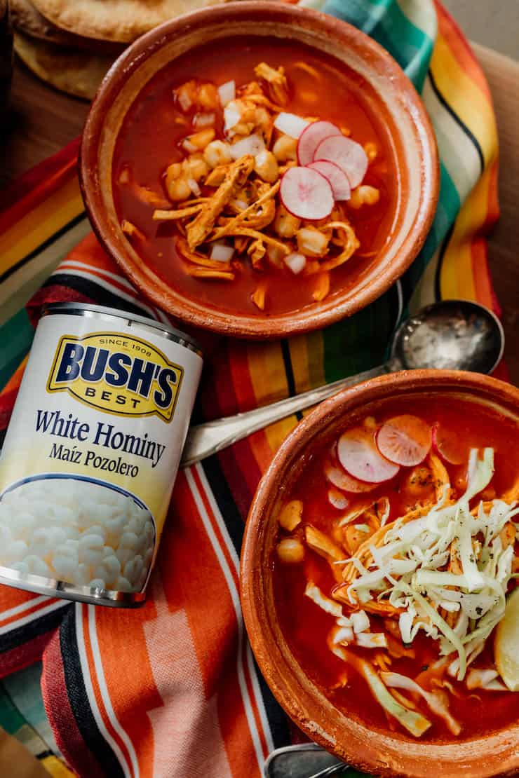 final overhead hero shot of red chicken pozole in terra cotta bowls with silver spoons and a can of bush's white hominy
