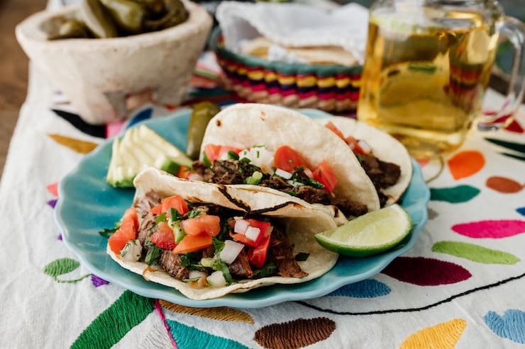 three slow cooker brisket tacos Suadero on a turquoise plate topped with pico de gallo and with a wedge of lime, sliced avocado and escabeche and beer on the side