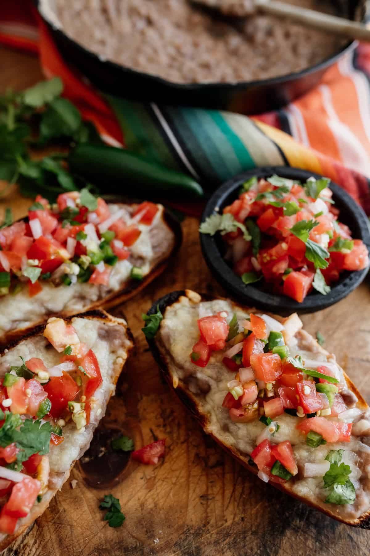 three molletes with refried beans, melted cheese and pico de gallo on a wooden cutting board with a black stone bowl holding more pico.