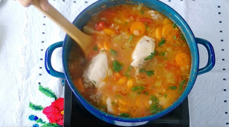 hand holding wooden spoon stirring together ingredients from sauté pan and chicken broth mixture for caldo de pollo