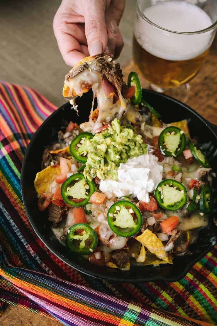 hand pulling a brisket nacho out of a cast iron skillet on a wood table with a pint of beer