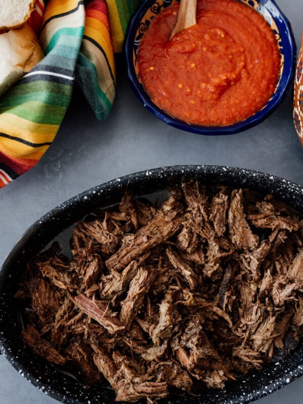 Shredded beef brisket in a black speckled dish next to a bowl of red salsa and bolillo rolls wrapped in a colorful Mexican servilleta.