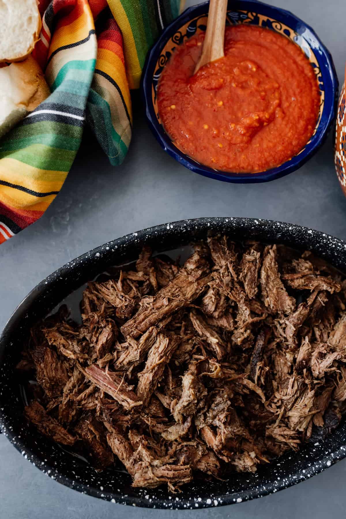 Shredded beef brisket in a black speckled dish next to a bowl of red salsa and bolillo rolls wrapped in a colorful Mexican servilleta.