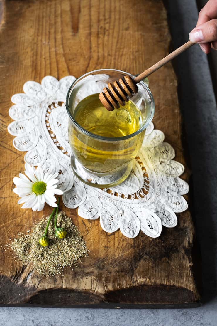 Té de manzanilla (Chamomile Tea) in a clear glass cup with a honey stick. The glass is on a vintage doily on a wood surface
