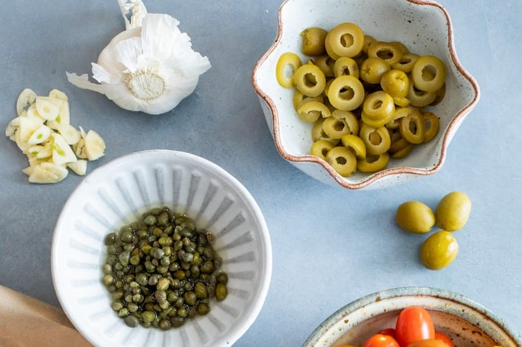bowls of capers, olives and tomatoes on a grey surface