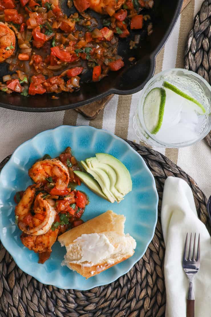 turquoise plate with camarones a la mexicana, sliced avocado and crusty bread topped with butter next to a pan full of mexican ranchero style shrimp