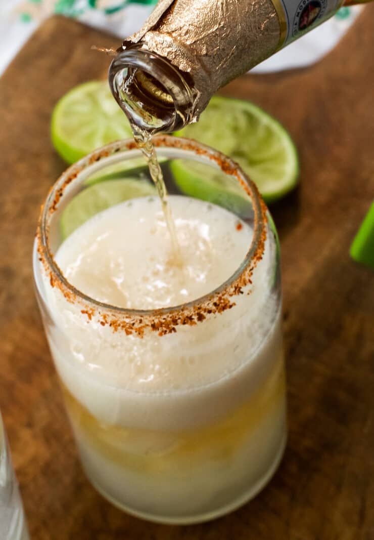 close up shot of Modelo beer being poured into a glass with ice and lime juice that has been rimmed with chili lime salt for a chelada