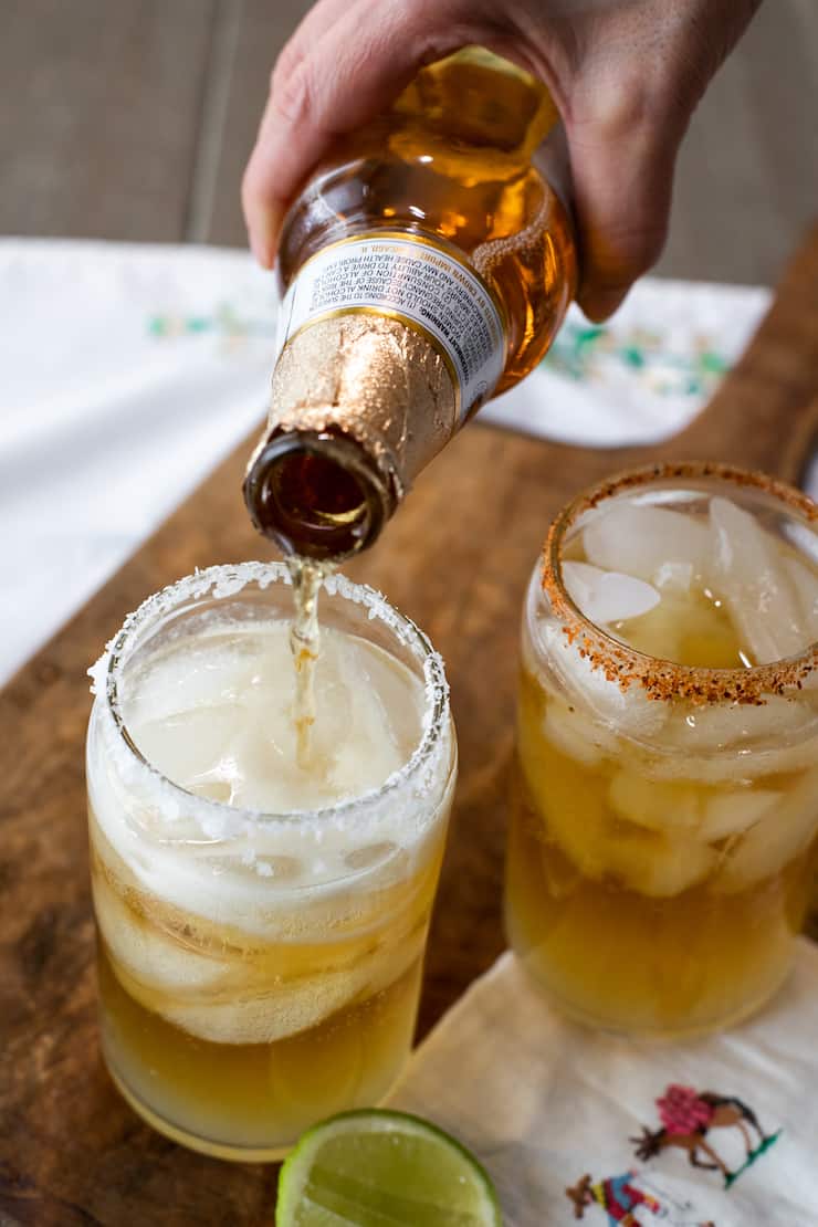 hand pouring modelo beer into salt rimmed glass with ice and lime juice