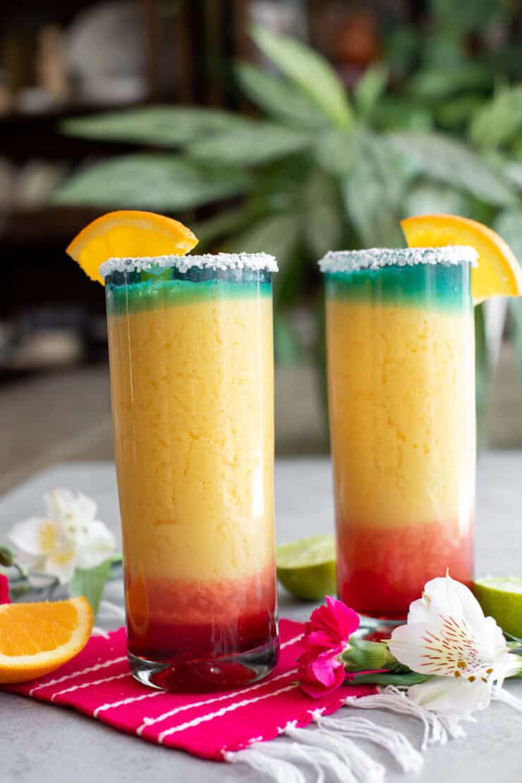 sideways shot of two tall collins glasses filled with frozen sunrise margaritas with a tropical plant background