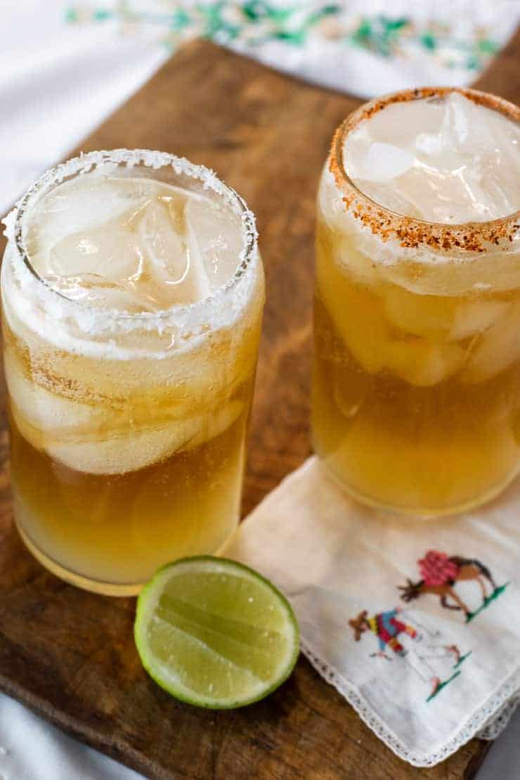 two chelada beer cocktails in clear glasses with salted rims on a wooden cutting board