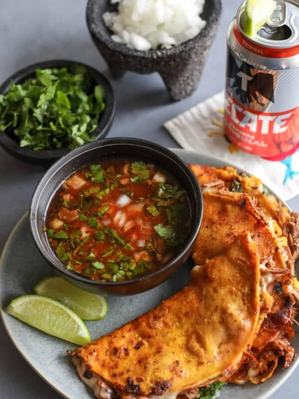 three quesabirria tacos on a grey plate with a side of birria dipping sauce and fresh lime wedges.