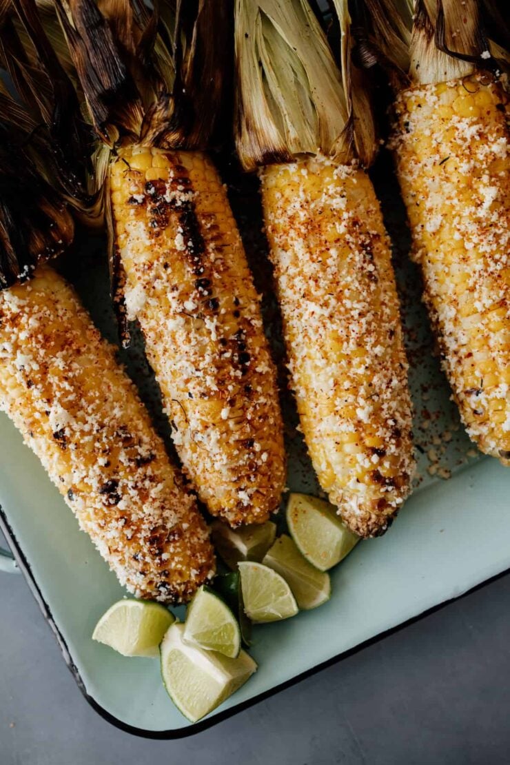4 cobs of corn that have been turned into elote on a teal rectangular serving dish with lime wedges