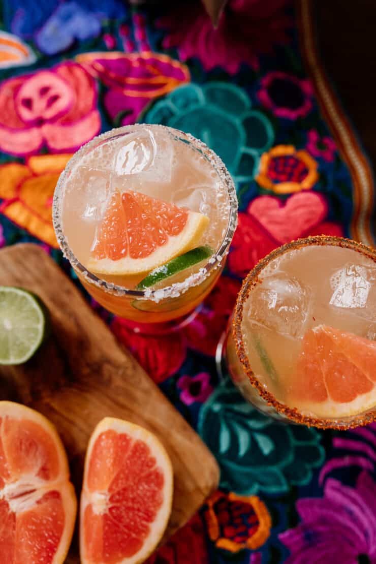 top down shot of two cocktail glasses prepped with Tajin and Salt with reposado tequila next to fresh grapefruit and lime wedges on a colorful Mexican flowered textile