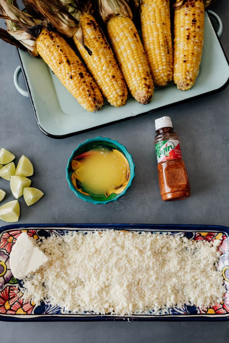 overhead shot of grilled corn on the cob with the fixings for making it into elote laid out on a grey table