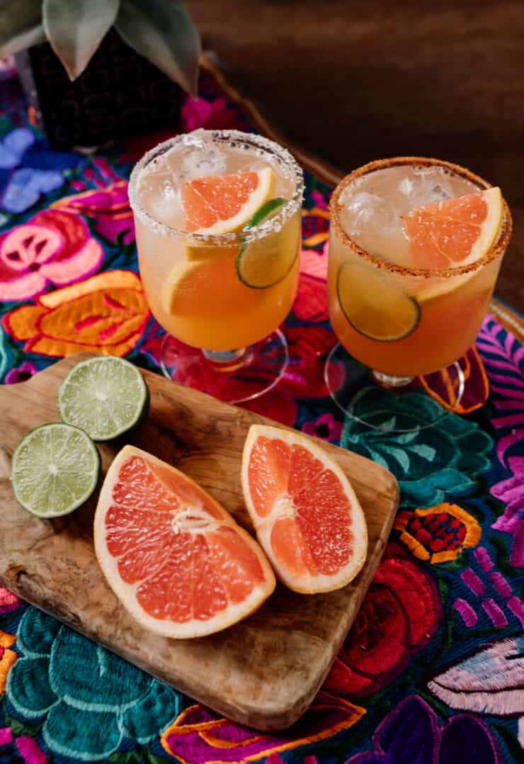 two cocktail glasses filled with reposado palomas next to fresh grapefruit and lime wedges on a colorful Mexican flowered textile