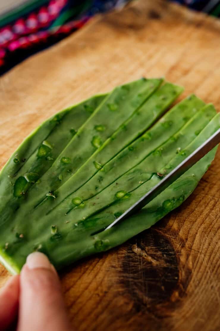 process shot - slicing nopales on a wooden cutting board in a fan shape where the base is still connected but long strips have been cut.