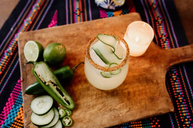 cucumber margarita with a Tajin rim and fresh jalapeno, and cucumber and limes next to drink