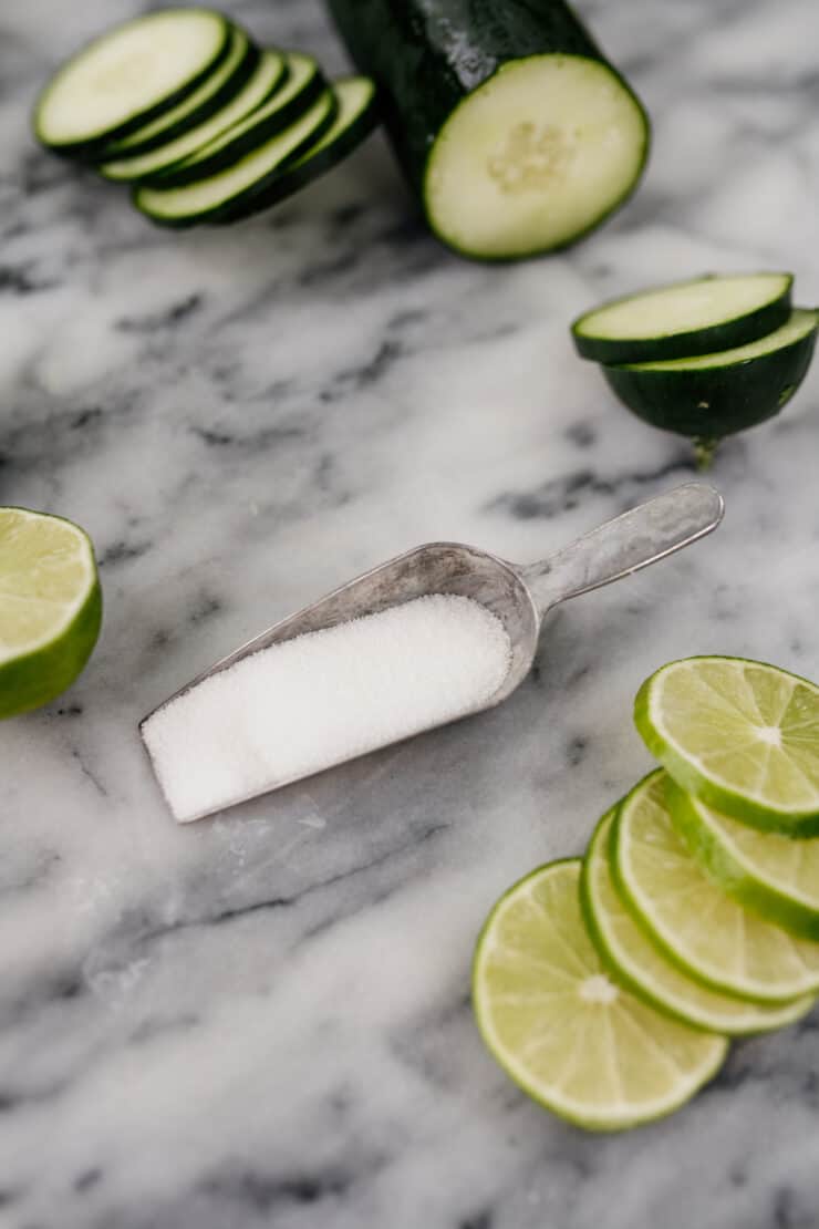 sliced cucumbers and limes on a marble tabletop with a silver scoop of sugar