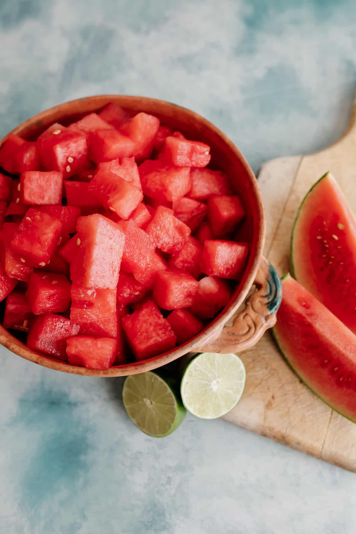 overhead shot of watermelon cubes in a terracotta bowl with a halved lime and some watermelon wedges on the side.