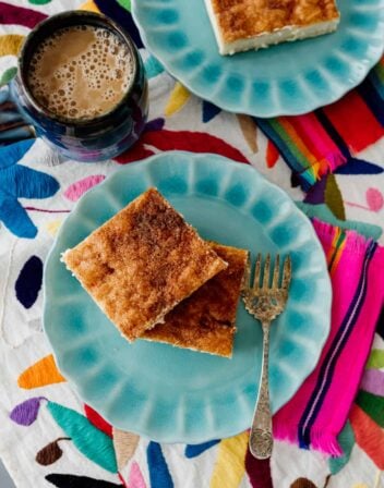 Sopapilla Cheesecake Bars on teal plates with a vintage dessert fork on colorful textiles with a cup of coffee.