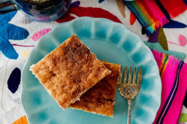 Sopapilla Cheesecake Bars on teal plates with a vintage dessert fork on colorful textiles with a cup of coffee.