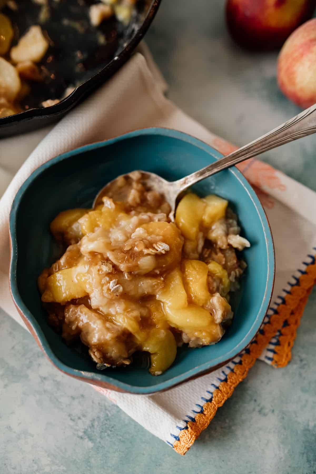 baked peach crisp in a turquoise bowl on an embroidered napkin with a spoon