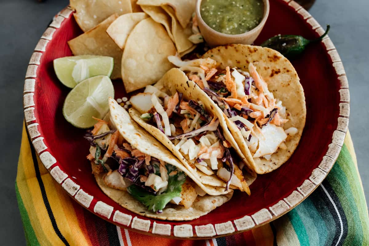 three cod fish tacos with coleslaw in a red plate and chips and salsa on the side.