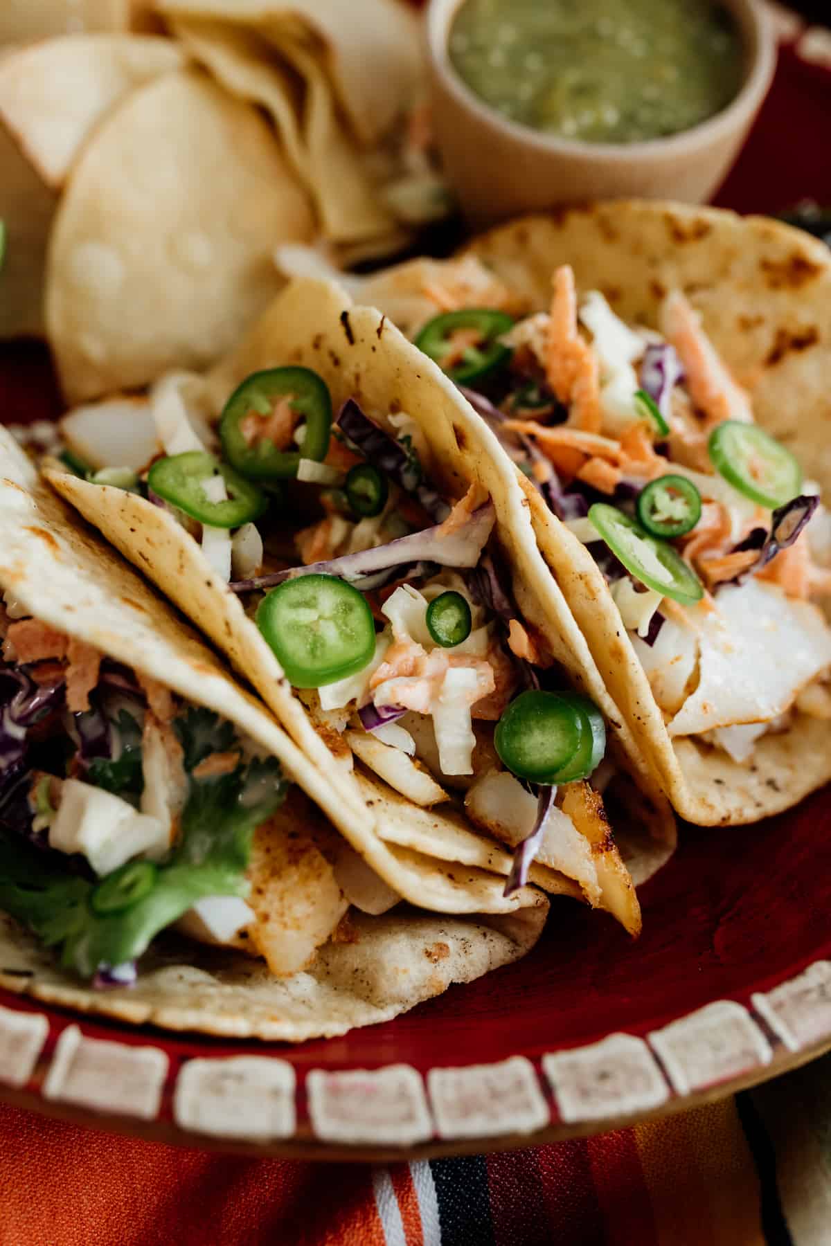 close up photo of cod fish tacos topped with slices of jalapeño with chips and salsa in the background.