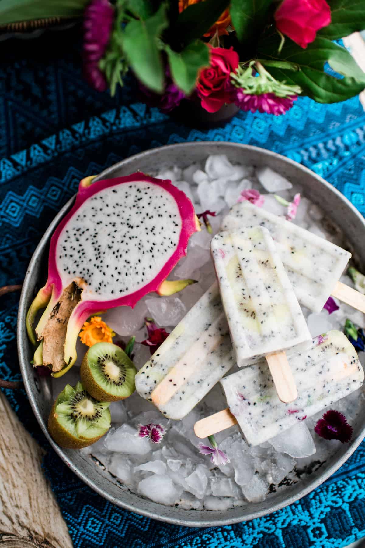 kiwi and dragron fruit paletas on ice in a silver metal tray with a blue runner underneath and flowers in the background 