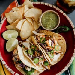 overhead shot of a round red platter with three cod fish tacos, a side of tortilla chips, salsa verde and sliced limes