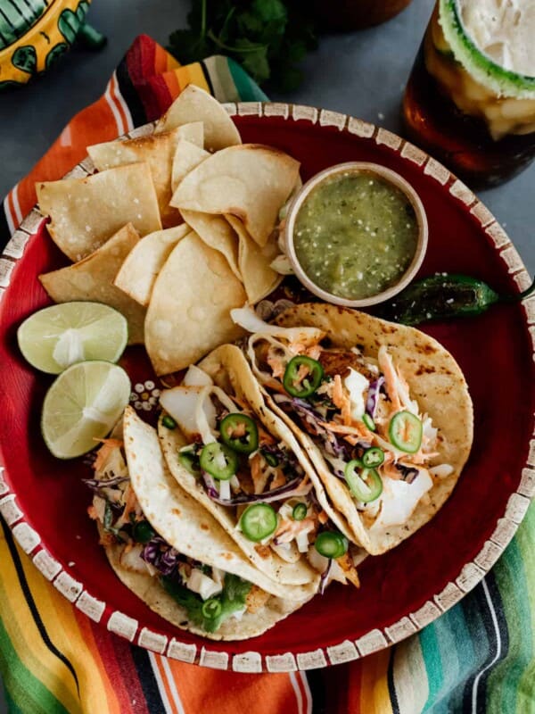 overhead shot of a round red platter with three cod fish tacos, a side of tortilla chips, salsa verde and sliced limes