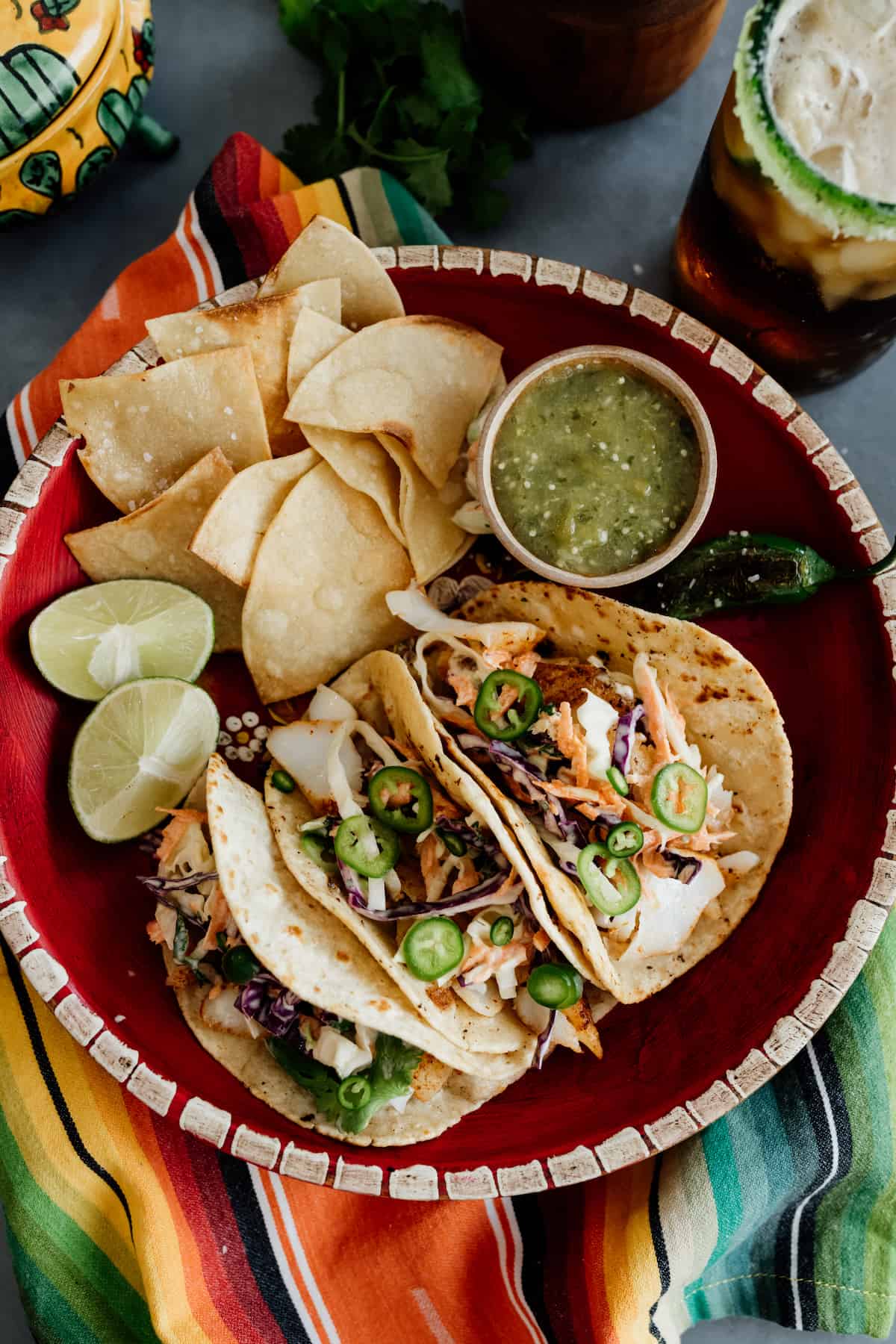 Cod fish tacos with slaw in a red bowl with a side of tortilla chips and salsa verde and a dark beer with a colorful serape napkin.