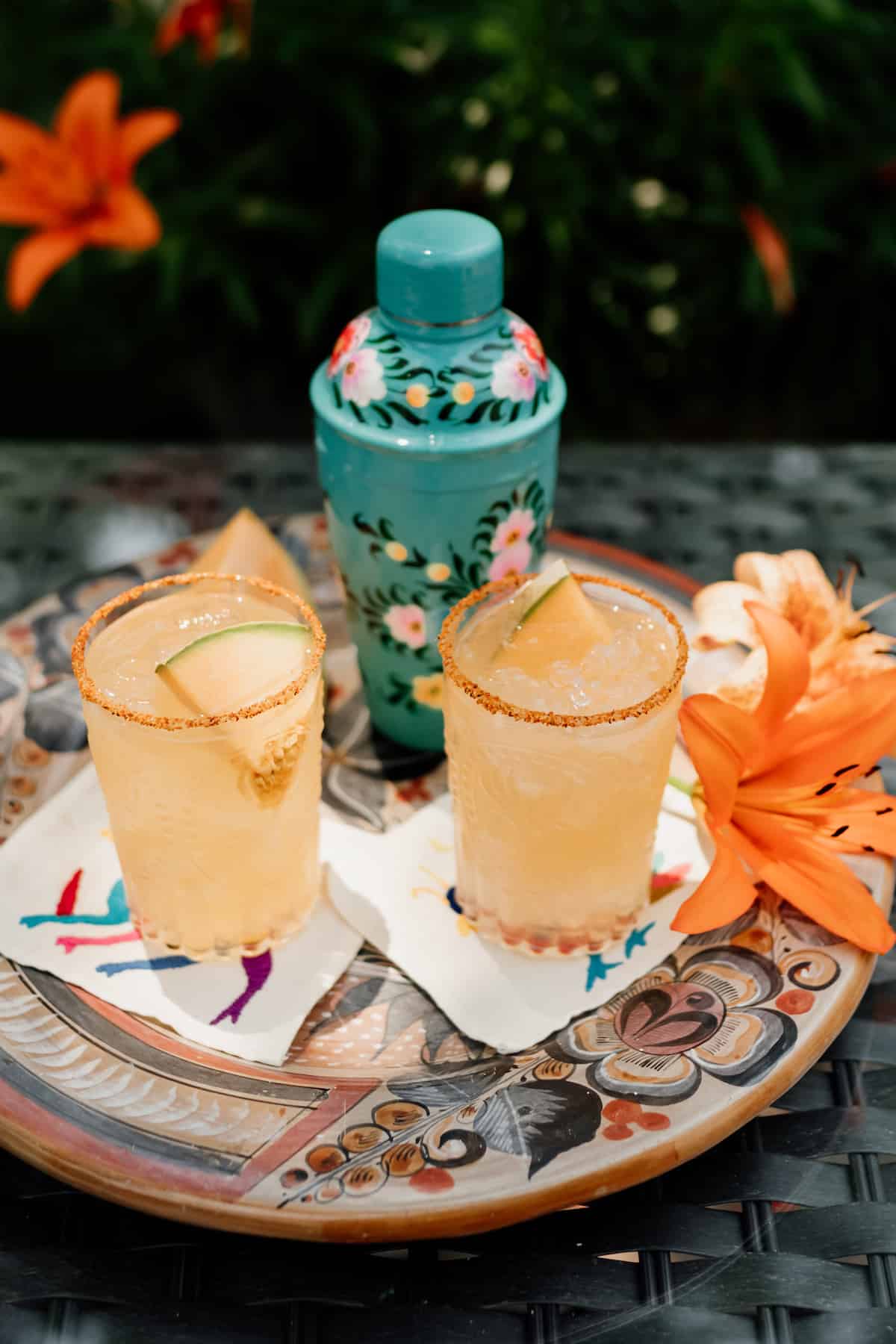 two glasses of Cantaloupe Melon Margaritas rimmed with Tajin and Lilly flowers on the side and cocktail shaker in the background 