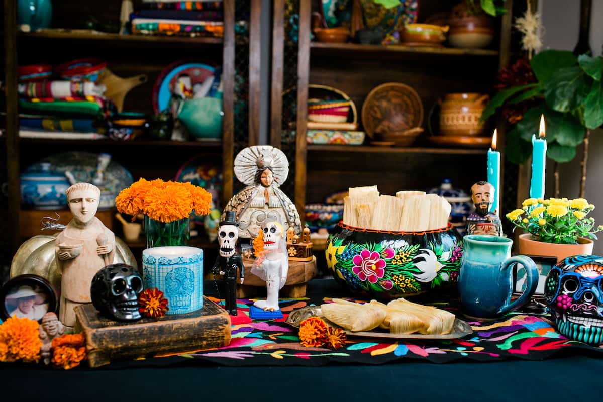 a mini alter for Dia de los Muertos adorned with candles, marigolds, figurines, and pumpkin spice tamales