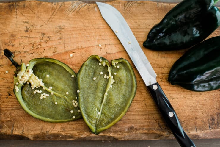two fresh poblano peppers on a wooden board and one roasted on a cast iron skillet 