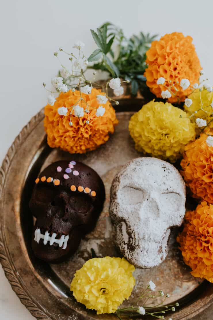 Day of the Dead Mexican Chocolate Skull Cakes—one dusted with powdered sugar and one decorated with chocolate—surrounded by baby’s breath and fresh marigold flowers.