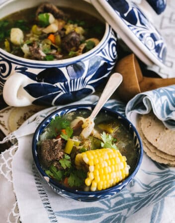blue and white Mexican soup tureen filled with oxtail soup and a stack of corn tortillas and a wooden ladle.