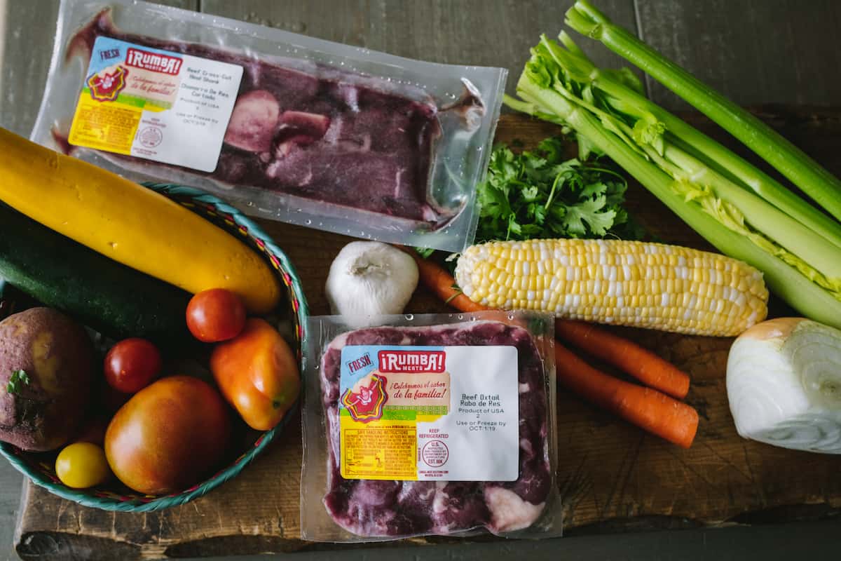 ingredients to make oxtail soup on a cutting board corn, celery, garlic, zucchini, tomatoes, beef, potatoes, carrots