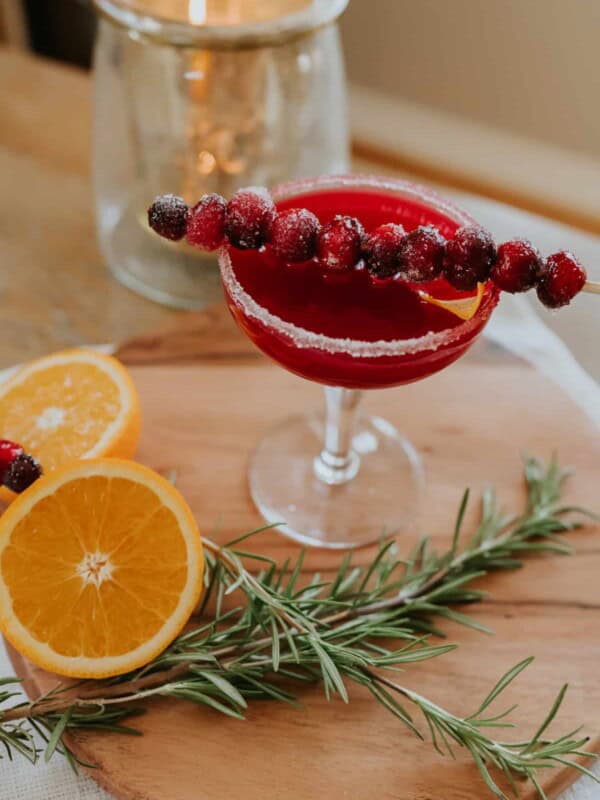 A festive cranberry margarita served with a skewer of sugared cranberries and a sugared rim in a martini class on a wooden cutting board with rosemary and orange slices as garnish. The perfect Christmas cocktail or Thanksgiving drink!